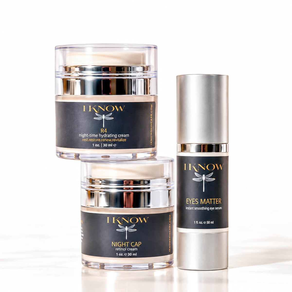 Three skincare products from I Know Beauty arranged on a white reflective surface: a stacked pair of clear jars with black labels and silver lids, and one tall silver cylindrical bottle labeled Eyes Matter.