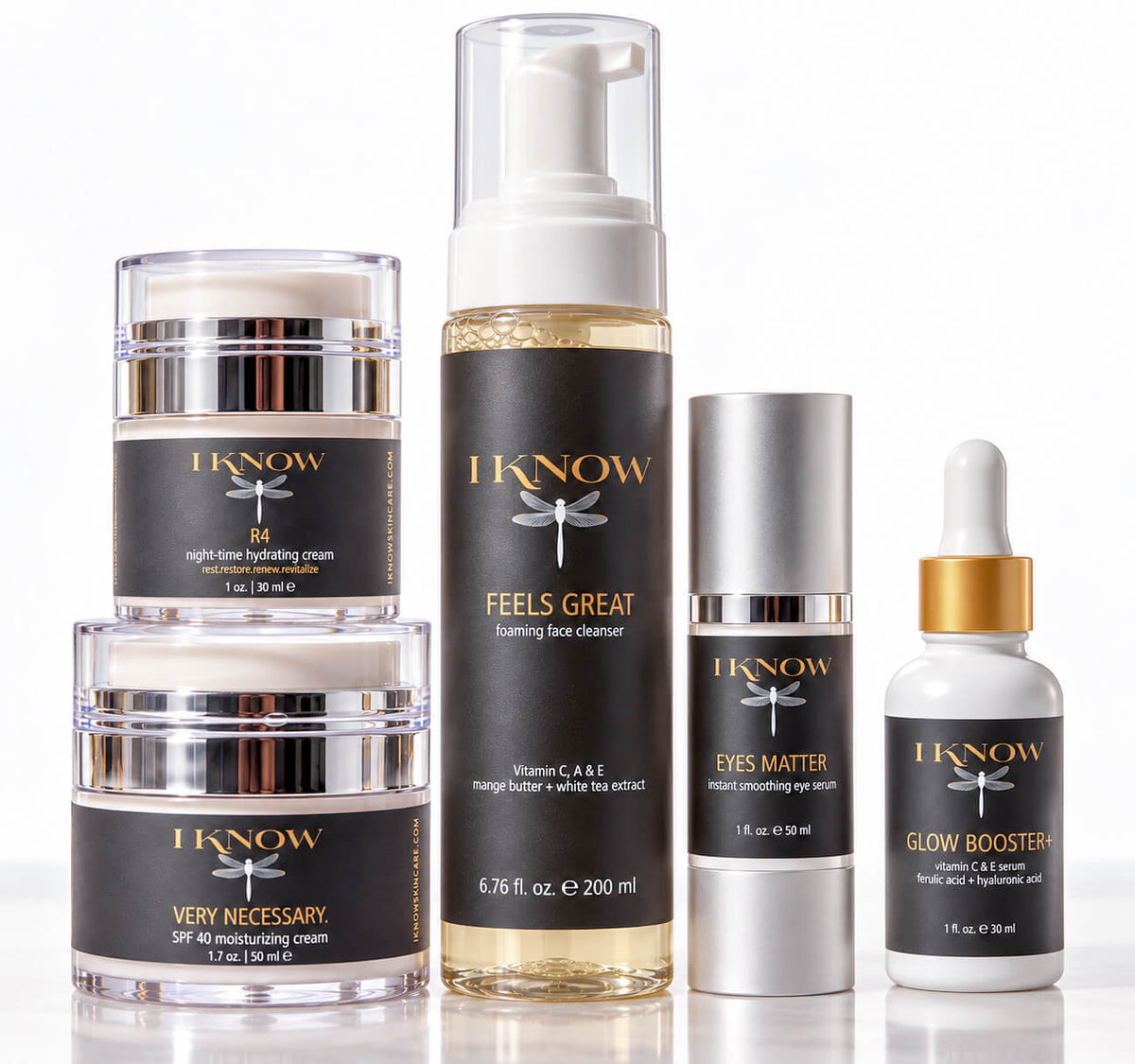A five-piece skincare set from IKnow Cosmetics arranged on a reflective white surface. The set includes two stacked jars of night-time hydrating cream, a tall bottle of foaming face cleanser, a bottle of eye cream, and a dropper bottle of glow booster, all featuring black labels with gold lettering and dragonfly logos.