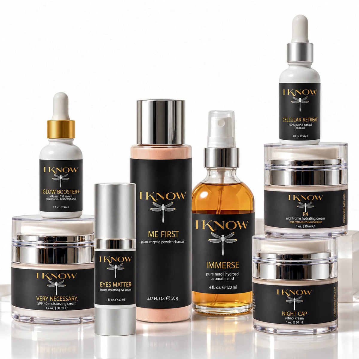 A collection of IKNOW skincare products arranged on a reflective white surface against a white background, including bottles, jars, and a pump bottle with black labels and silver and gold caps.